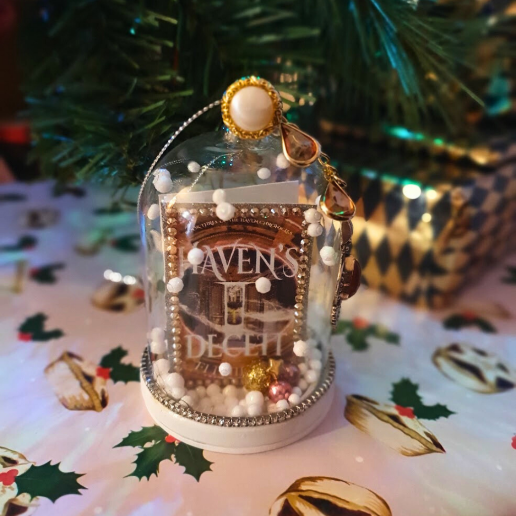 a glass dome Christmas Bauble with a tiny version of Haven's Deceit by Fi Phillips in side against a backdrop of a Christmas tree and presents